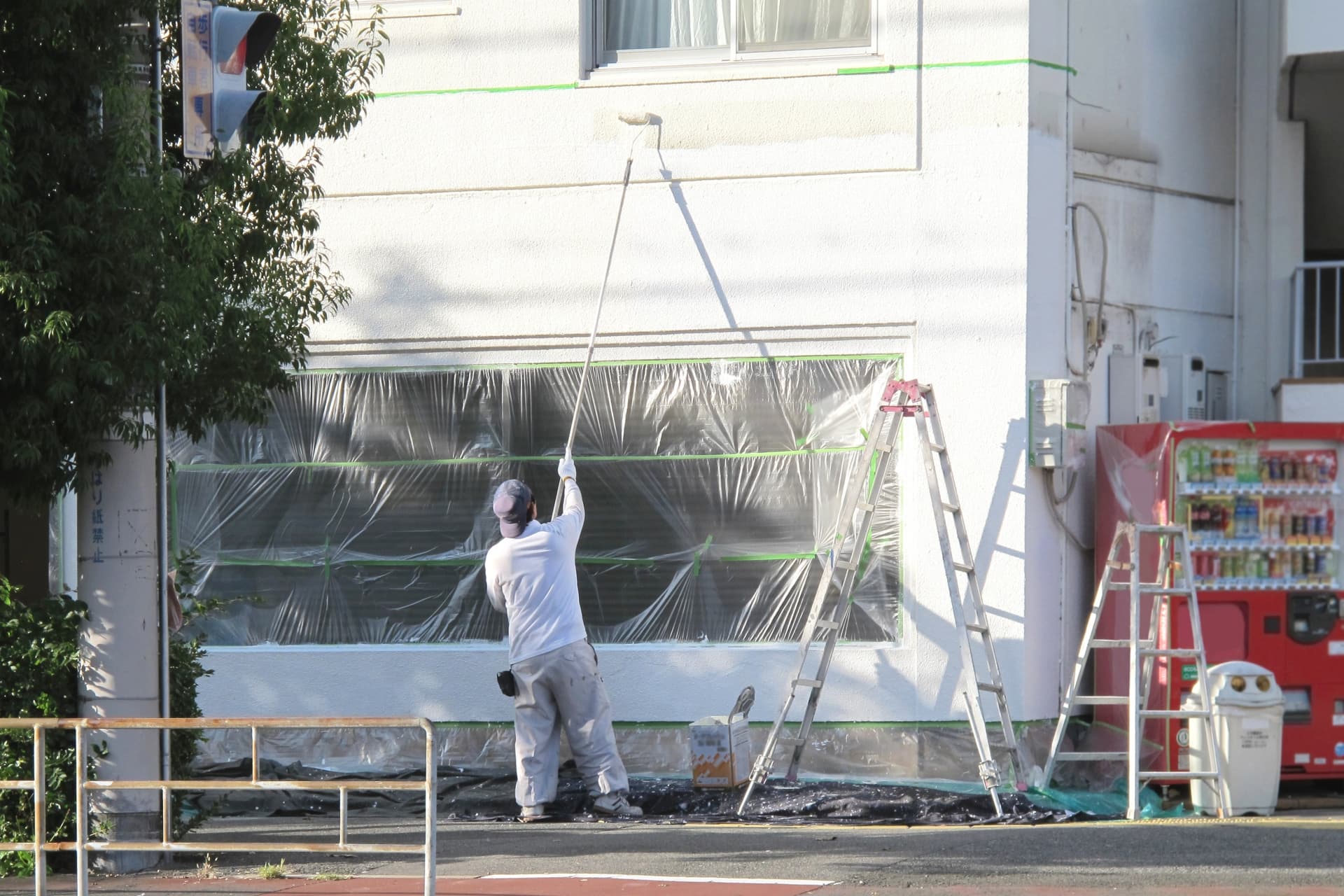 窓が養生された状態で建物の外壁の上の部分を塗装している様子